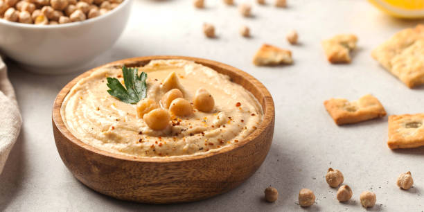 A bowl of hummus garnished with chickpeas and parsley, surrounded by crackers and chickpeas on a light surface.