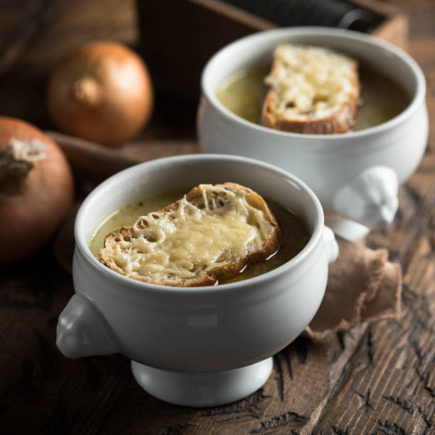 Two bowls of onion soup topped with melted cheese and bread slices, with onions in the background.