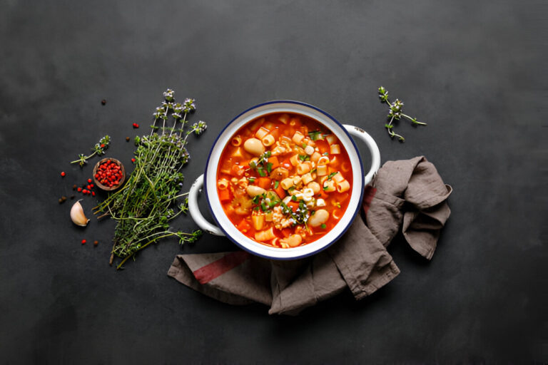 A bowl of colorful vegetable soup with pasta, surrounded by fresh herbs and spices.