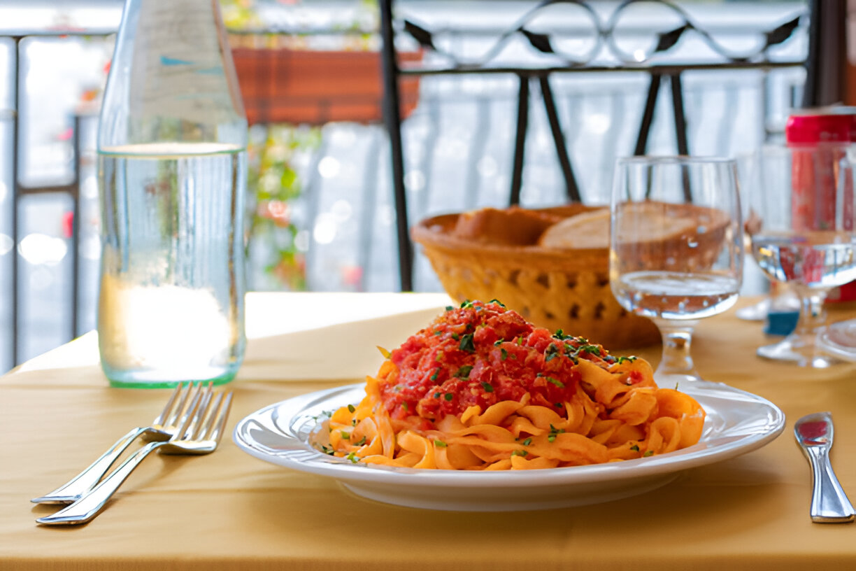 A plate of pasta with tomato sauce and herbs, presented on a table with a full glass of water, a bottle of water, and a basket of bread in the background.