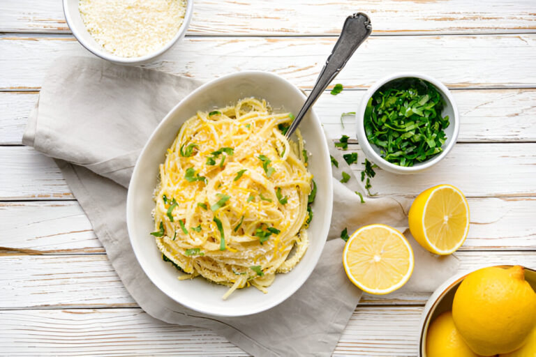 A bowl of linguine pasta garnished with green herbs, surrounded by fresh lemons and a bowl of grated cheese.