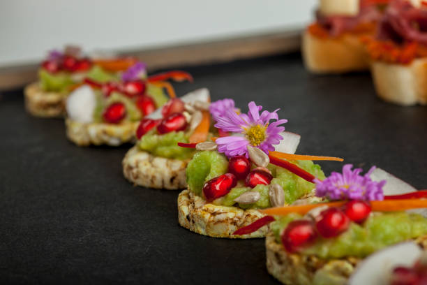 Gourmet canapés with colorful toppings including pomegranate seeds, green spread, and flowers, arranged on a dark slate serving board.