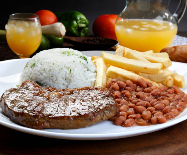 A plated meal featuring a juicy steak, white rice with green herbs, fried potatoes, and pinto beans, accompanied by a glass of orange beverage.