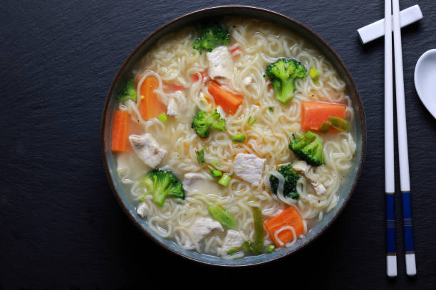 A bowl of chicken noodle soup with broccoli, carrots, and strips of chicken on a dark surface.