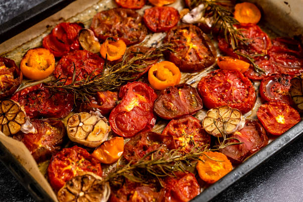 A baking tray filled with roasted tomatoes, orange peppers, and garlic, garnished with sprigs of rosemary.