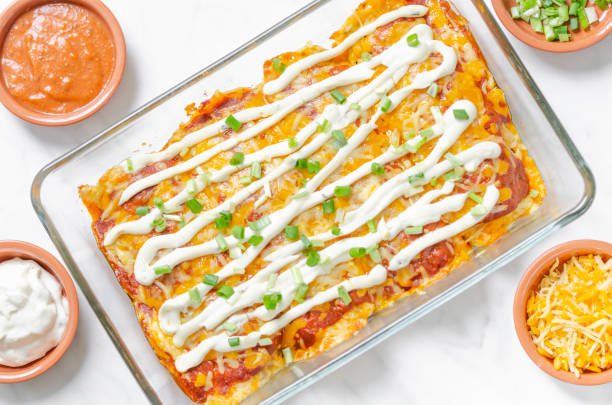 A rectangular glass dish filled with enchiladas topped with sour cream and sliced green onions, surrounded by small bowls of red sauce and shredded cheese.