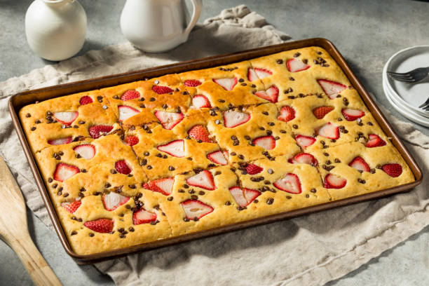 A freshly baked sheet cake topped with sliced strawberries and chocolate chips on a rustic table.