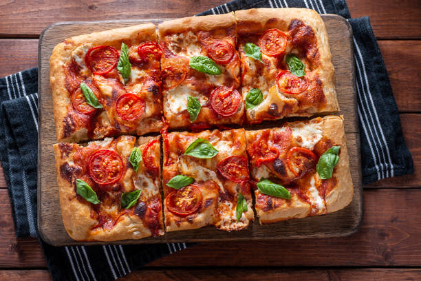 A freshly baked square pizza topped with sliced tomatoes and basil leaves, served on a wooden board.