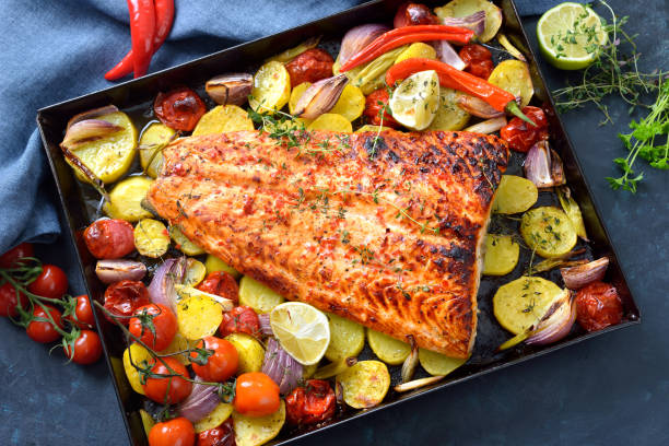 A beautifully roasted salmon fillet surrounded by vibrant vegetables including potatoes, tomatoes, and colorful peppers on a baking tray.