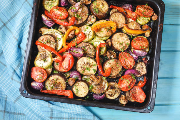 A tray of roasted vegetables including eggplant, bell peppers, tomatoes, and mushrooms, garnished with herbs, set on a blue tablecloth.