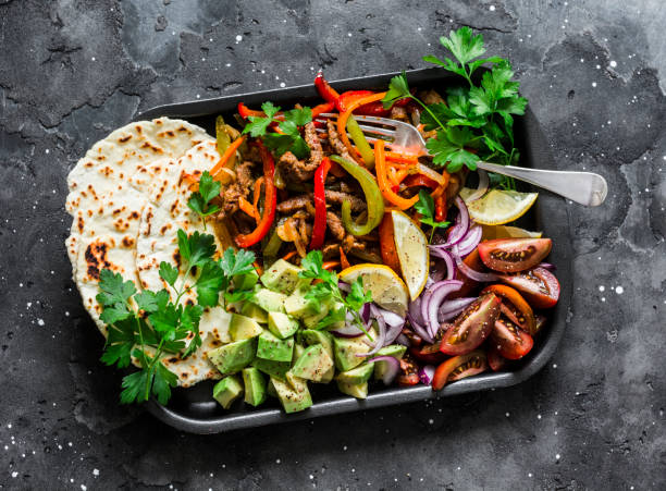 A colorful mixed platter featuring grilled vegetables, sliced avocado, tomatoes, and flatbreads on a dark textured background.