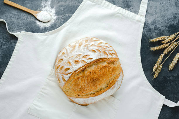 Freshly baked round bread on a white apron with a wooden spoon and wheat stalks nearby.