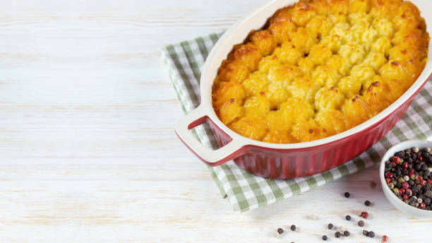 A freshly baked dish in a red ceramic casserole with a golden-brown crust, resting on a checkered napkin with a small bowl of mixed peppercorns nearby.