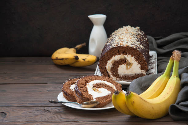 Chocolate roll cake with cream filling, sliced on a plate, alongside fresh bananas and a white jug.