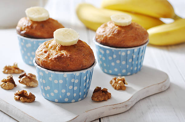 Three banana muffins with walnuts on a wooden board, decorated with banana slices, and served with bananas in the background.