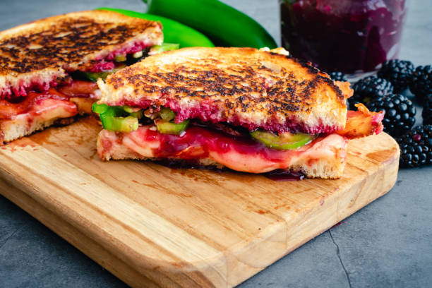 A close-up of a grilled sandwich with layers of peanut butter, jelly, and sliced green peppers, served on a wooden cutting board.