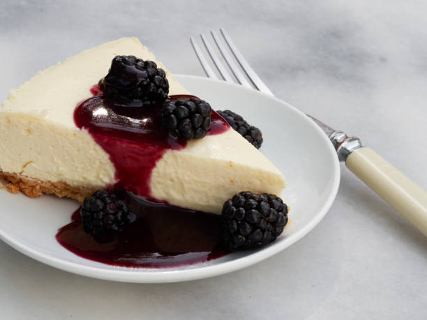 A slice of cheesecake topped with blackberries and a berry sauce on a white plate with a fork beside it.