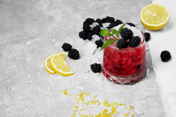 A refreshing cocktail garnished with blackberries and mint leaves, served with lemon slices on a textured gray background.