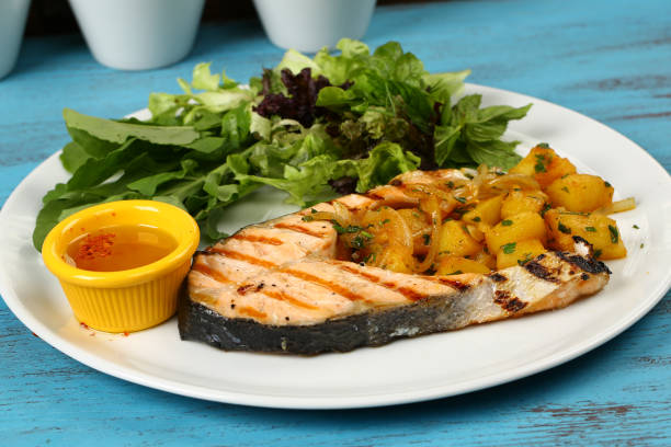 A plate of grilled salmon with sautéed vegetables and a side salad, served with a small cup of sauce.