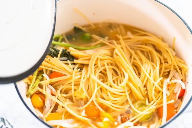 A pot of spaghetti noodles with mixed vegetables in a broth, partially covered by a lid.