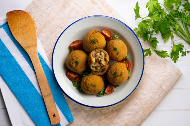 A bowl of golden-brown croquettes garnished with fresh parsley and cherry tomatoes, served with a wooden spoon beside it on a textured surface.