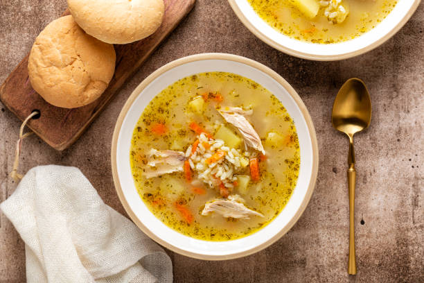 Two bowls of chicken soup with rice and vegetables placed beside a wooden board with rolls.