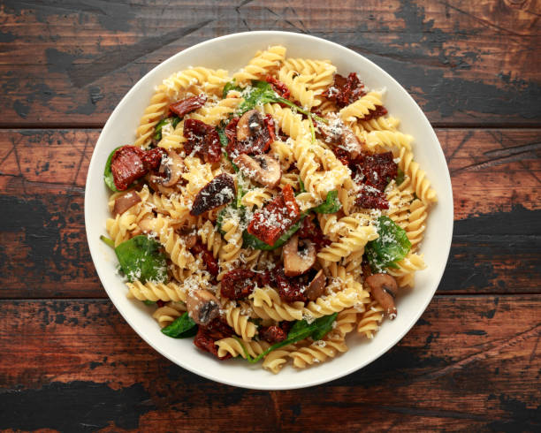 A plate of spiraled pasta topped with spinach, mushrooms, sun-dried tomatoes, and grated cheese on a wooden table.