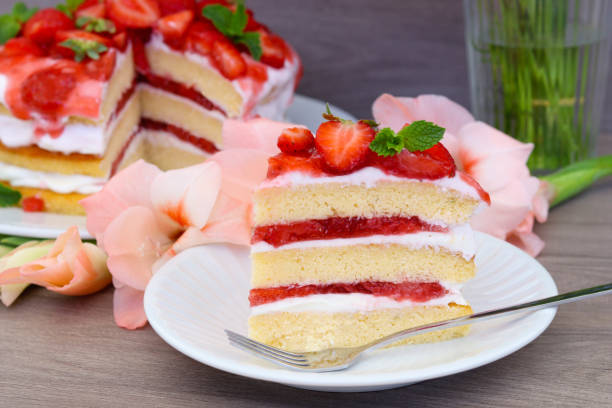 A slice of strawberry cake on a white plate with a fork, surrounded by pink flowers and a blurred background of a whole cake and a glass of water.