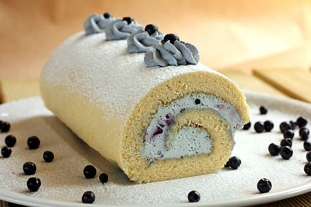 A rolled cake with cream filling, decorated with blue frosting and powdered sugar, placed on a white plate with black currants scattered around.