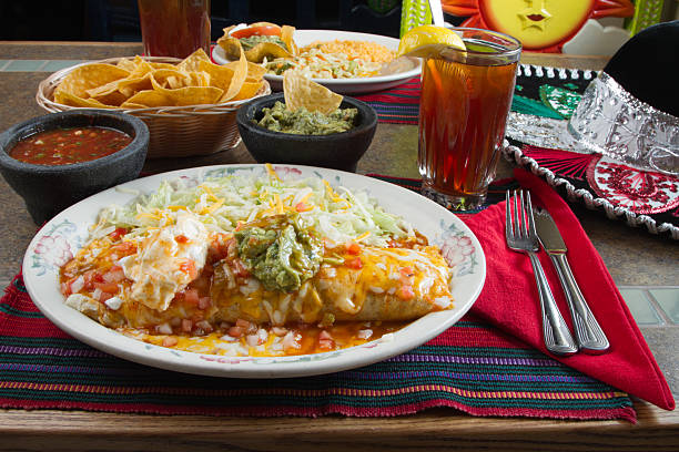 A colorful Mexican meal featuring a large enchilada topped with cheese, lettuce, and guacamole on a decorative plate, accompanied by tortilla chips, salsa, and a refreshing drink.