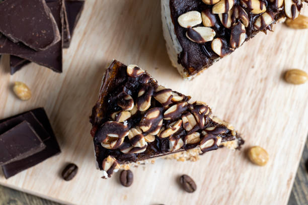 A slice of rich chocolate cake topped with nuts, served on a wooden board alongside chocolate pieces and coffee beans.