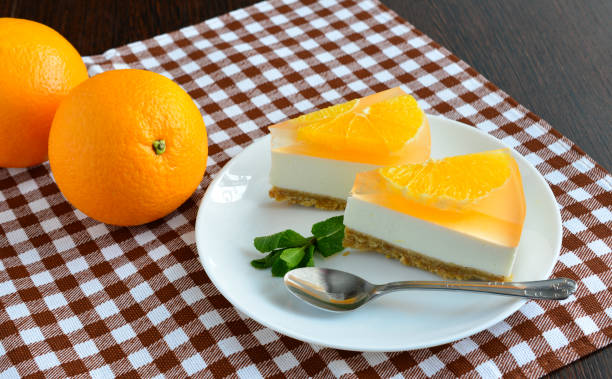 A plate of orange-flavored cheesecake slices garnished with orange segments, mint leaves, and a spoon, beside two whole oranges on a checkered tablecloth.