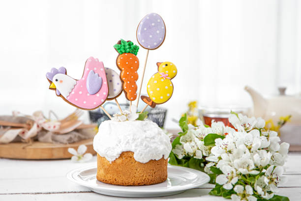 A festive cake topped with whimsical decorated cookies shaped like a chicken, carrot, and egg, surrounded by white flowers.