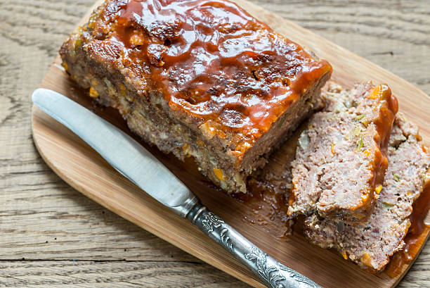 A sliced homemade meatloaf with a glossy glaze on a wooden cutting board, accompanied by a silver knife.