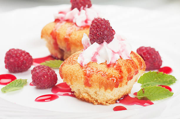 Delicious vanilla cupcakes topped with whipped cream and fresh raspberries, drizzled with raspberry sauce, arranged on a white plate.