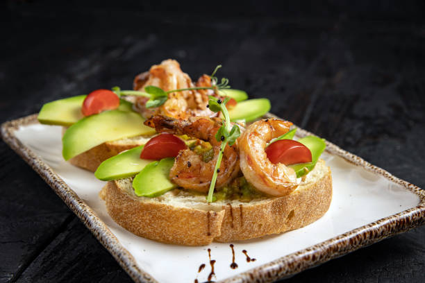 A beautifully plated dish featuring shrimp on toasted bread with avocado slices and cherry tomatoes, garnished with microgreens.