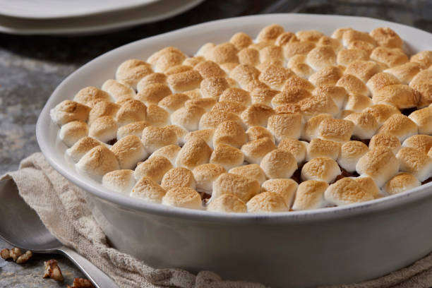 A close-up of a baked dish topped with golden-brown marshmallows, resting on a countertop with a napkin and walnuts nearby.