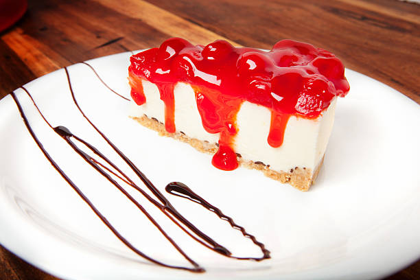 A slice of cheesecake topped with a bright red cherry sauce, served on a white plate with chocolate drizzle.