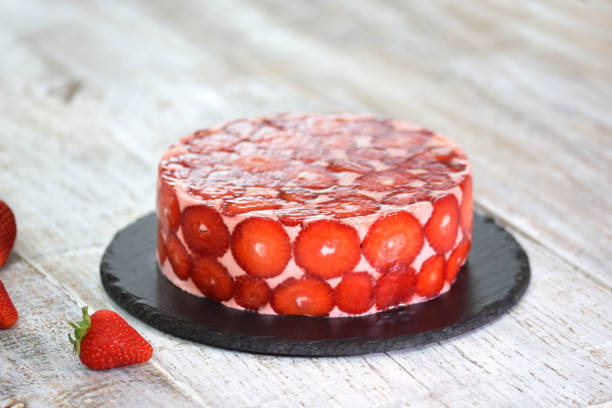 A round strawberry cake with a glossy red glaze on a black slate plate, surrounded by fresh strawberries