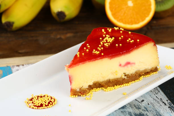 A slice of cheesecake topped with red gelatin and yellow sprinkles on a white plate, with bananas and an orange in the background.