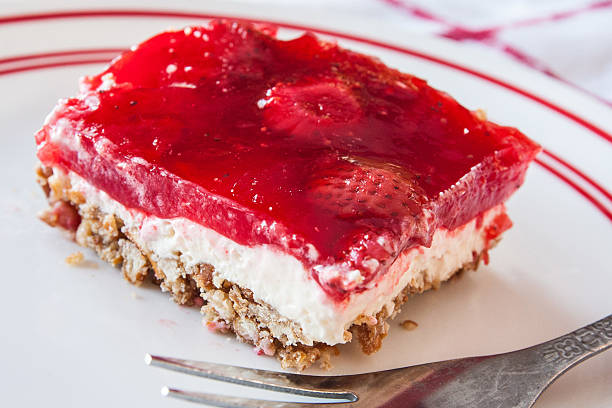 A slice of dessert with a red jelly topping on a cream layer, resting on a crumb crust, served on a white plate with a fork.