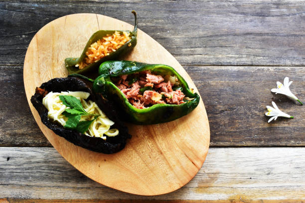 A wooden serving platter with three stuffed peppers: one green pepper filled with rice, one green pepper containing shredded meat, and one blackened pepper filled with cheese and herbs.