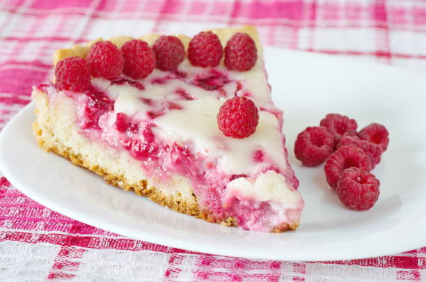 A slice of raspberry cheesecake on a white plate, garnished with fresh raspberries, sitting on a pink and white checkered tablecloth.