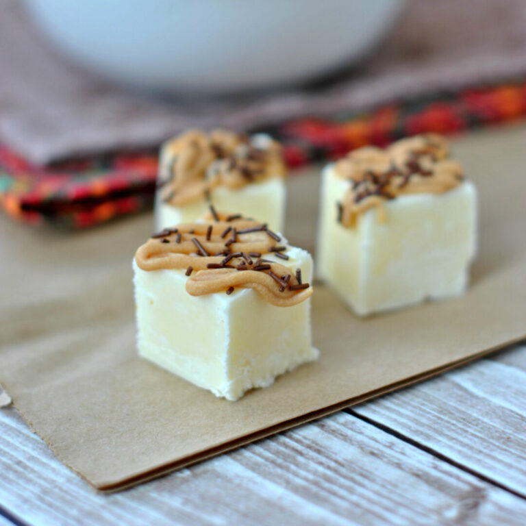 Three white fudge squares topped with peanut butter frosting and chocolate sprinkles on a piece of brown parchment paper.