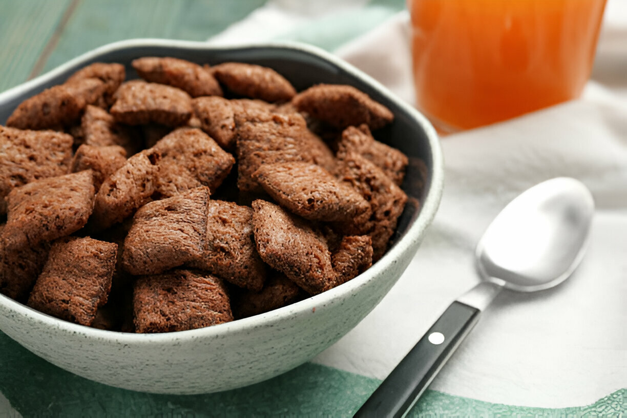 A bowl of chocolate-flavored square cereal pieces next to a spoon and a glass of orange juice.