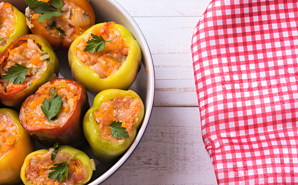 A close-up view of stuffed bell peppers filled with rice and meat, garnished with parsley, displayed in a round dish beside a red and white checkered cloth.