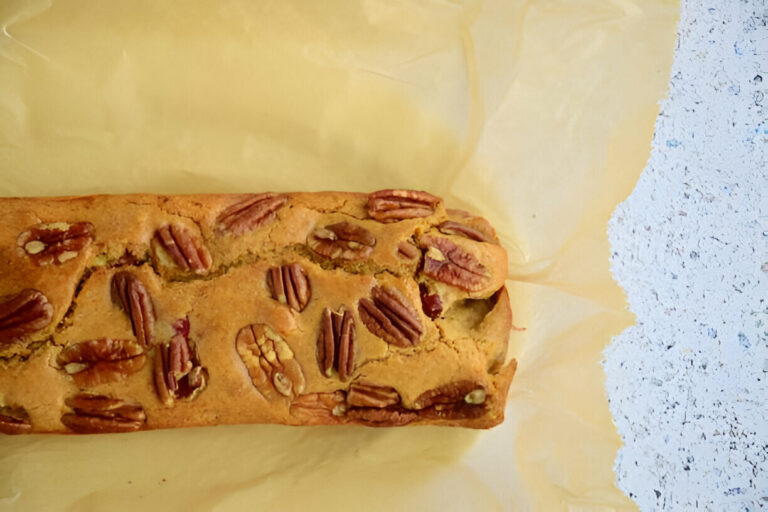 A freshly baked loaf of pecan bread with visible pecan halves on top, resting on yellow parchment paper.