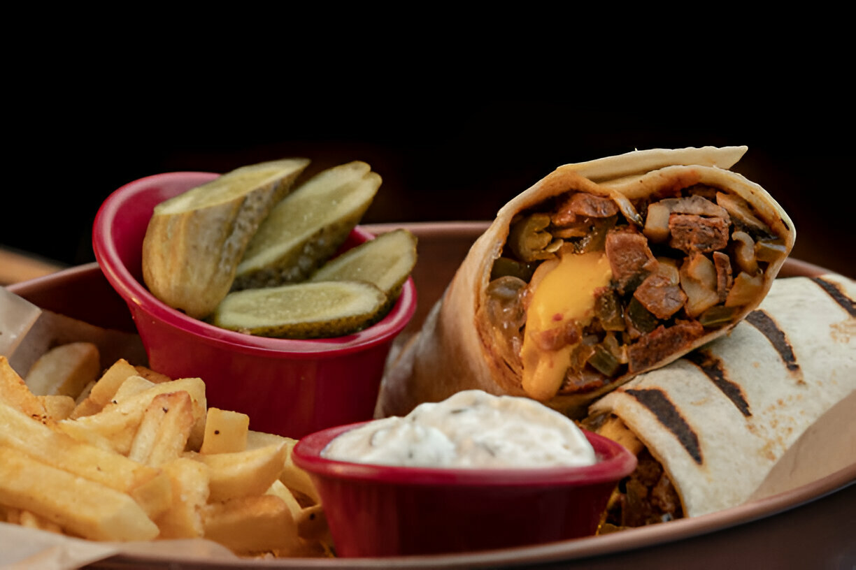 A plate featuring a burrito, French fries, pickles, and a small bowl of dipping sauce.