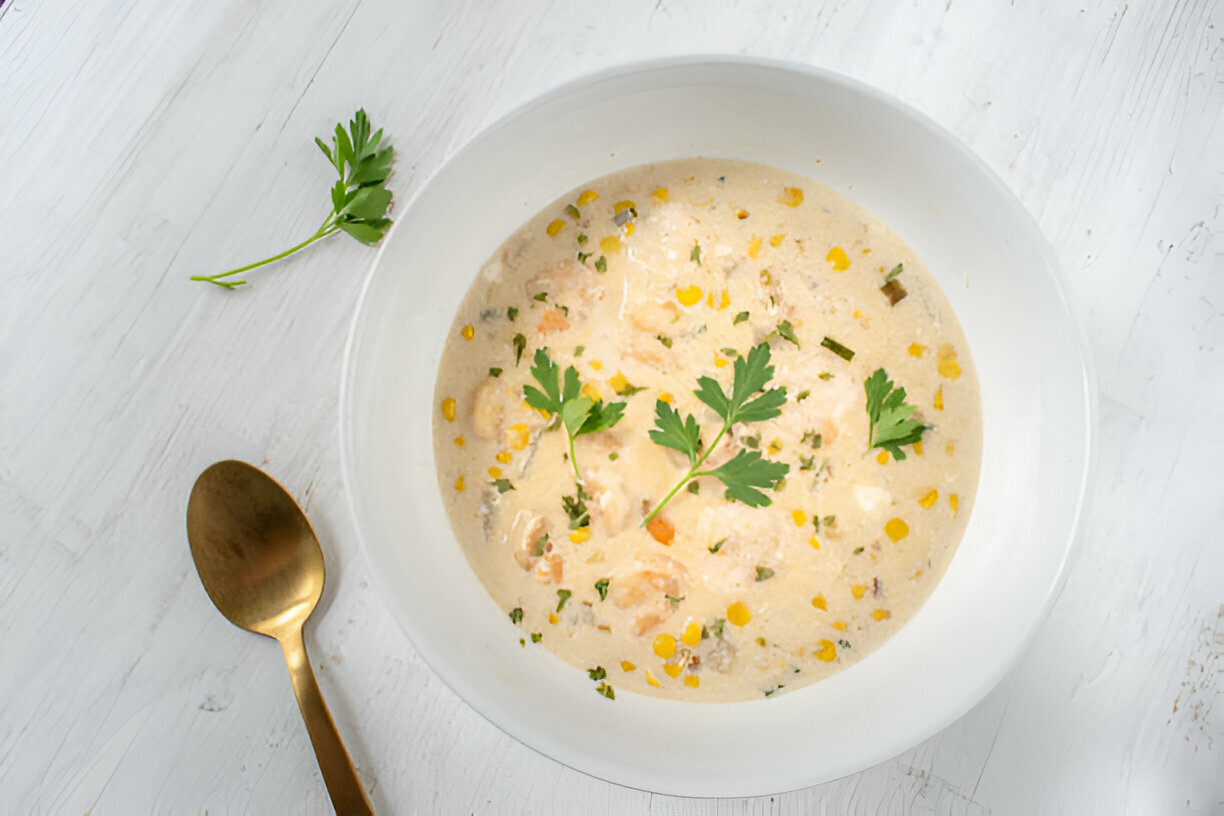 A creamy soup with shrimp and corn served in a white bowl surrounded by fresh herbs.