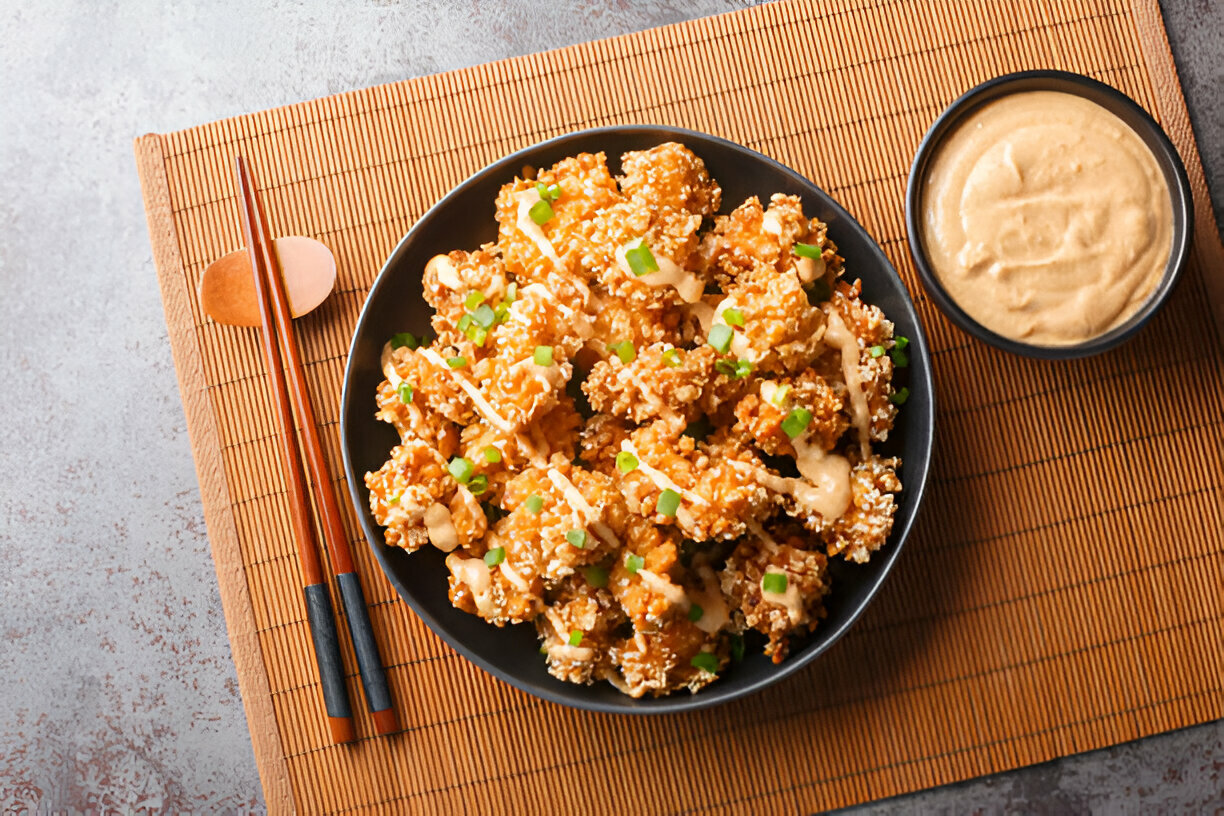 A bowl of crispy cauliflower topped with green onions and drizzled with sauce, served with a side of creamy dip on a bamboo mat.